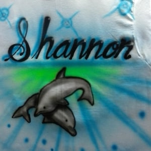 May include: White t-shirt with the name "Shannon" in black script, airbrushed with a blue and green background. Two gray dolphins are featured below the name, with blue starburst accents.