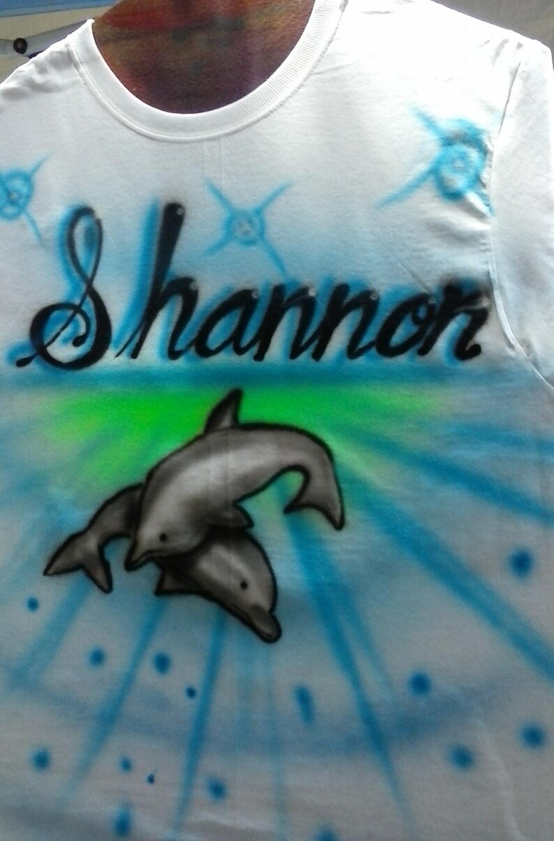May include: White t-shirt with the name "Shannon" in black script, airbrushed with a blue and green background. Two gray dolphins are featured below the name, with blue starburst accents.