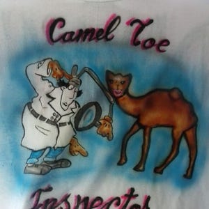 May include: White t-shirt with an airbrushed design featuring a cartoon camel and a detective character. The text "Camel Toe Inspector" is written in pink and black script. The design is set against a blue background.
