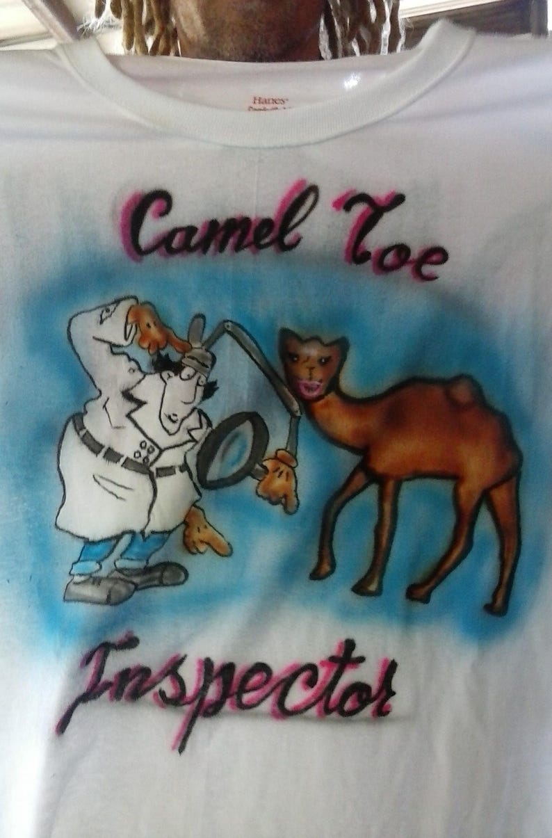 May include: White t-shirt with an airbrushed design featuring a cartoon camel and a detective character. The text "Camel Toe Inspector" is written in pink and black script. The design is set against a blue background.