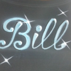 May include: Black garment with the name "Bill" in light blue, cursive lettering. The name is accented with sparkling starbursts. The design is airbrushed on the fabric.