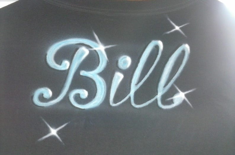 May include: Black garment with the name "Bill" in light blue, cursive lettering. The name is accented with sparkling starbursts. The design is airbrushed on the fabric.