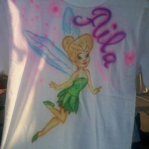May include: White t-shirt featuring a cartoon image of a fairy in a green dress with blue wings. The name "Aila" is written in purple script. Pink and blue airbrush accents surround the fairy.