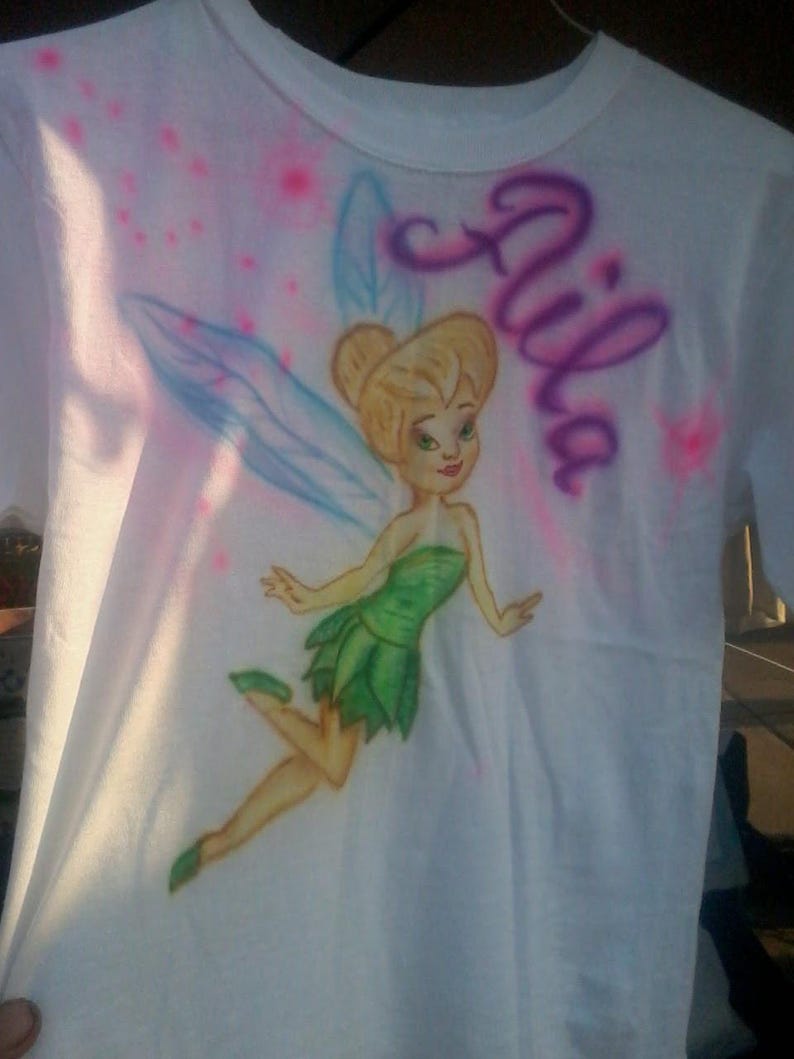 May include: White t-shirt featuring a cartoon image of a fairy in a green dress with blue wings. The name "Aila" is written in purple script. Pink and blue airbrush accents surround the fairy.