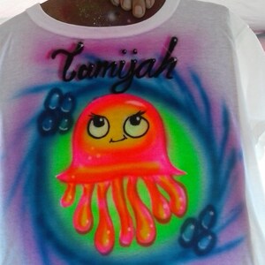 May include: White t-shirt with a colorful airbrushed design. The design features a cartoon jellyfish in shades of pink, orange, and yellow, set against a green and blue swirl. The name "Tamiyah" is written above the jellyfish in black script.
