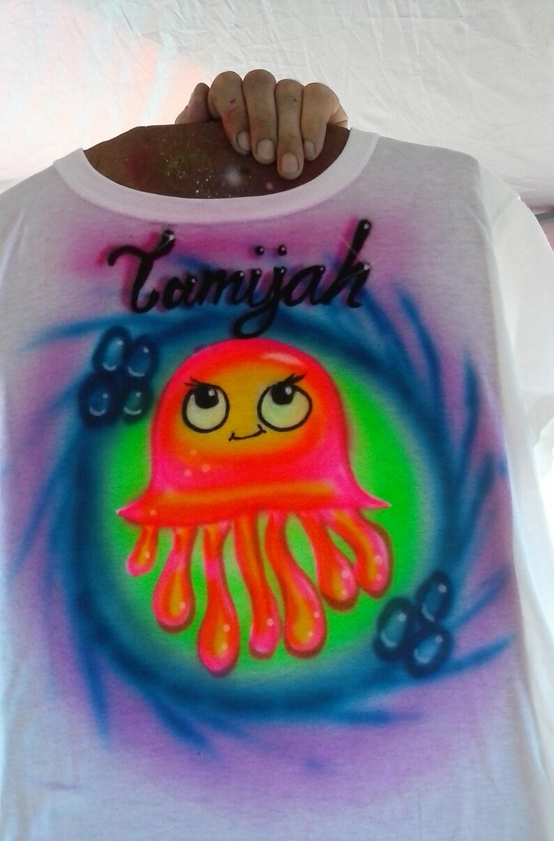 May include: White t-shirt with a colorful airbrushed design. The design features a cartoon jellyfish in shades of pink, orange, and yellow, set against a green and blue swirl. The name "Tamiyah" is written above the jellyfish in black script.