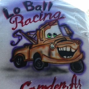 May include: White t-shirt featuring an airbrushed image of the character Tow Mater from the movie Cars. The design includes the text "Lo Ball Racing" and "Camden, Ar" in red and purple.