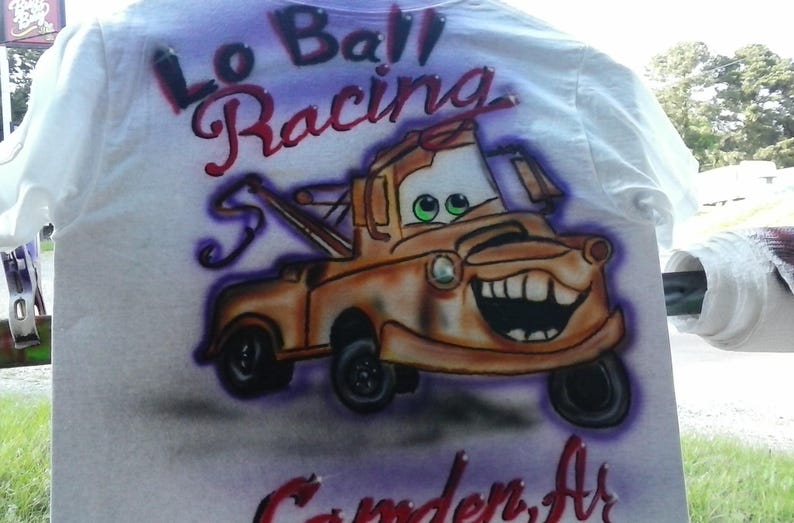 May include: White t-shirt featuring an airbrushed image of the character Tow Mater from the movie Cars. The design includes the text "Lo Ball Racing" and "Camden, Ar" in red and purple.