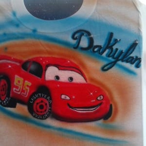 May include: White t-shirt featuring a red cartoon race car with the number 95 on the side. The car has a smiling face and is set against a blue and orange background. The name "Dakylan" is written above the car in cursive.