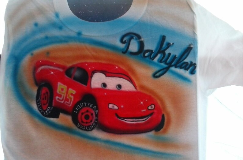 May include: White t-shirt featuring a red cartoon race car with the number 95 on the side. The car has a smiling face and is set against a blue and orange background. The name "Dakylan" is written above the car in cursive.