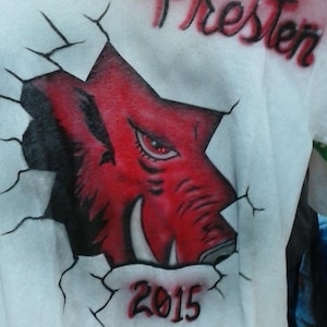 May include: White t-shirt featuring a red and black illustration of a Razorback hog breaking through a wall. The name "Presten" is arched above the design, with the year "2015" below. The design is airbrushed.