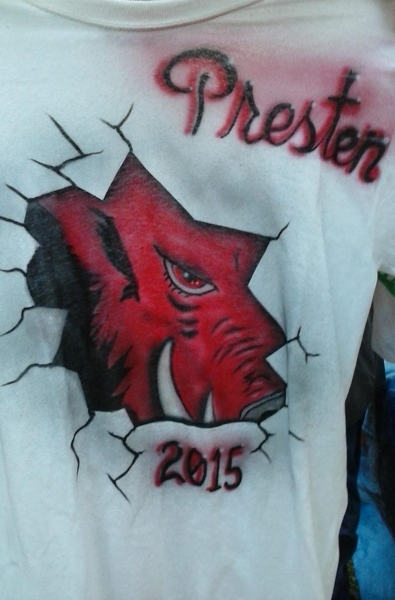 May include: White t-shirt featuring a red and black illustration of a Razorback hog breaking through a wall. The name "Presten" is arched above the design, with the year "2015" below. The design is airbrushed.