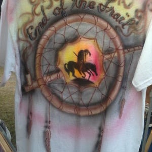May include: White t-shirt with an airbrushed design featuring a dreamcatcher, a Native American figure on horseback, and the text "End of the Trail". The design incorporates shades of brown, pink, yellow, and black.