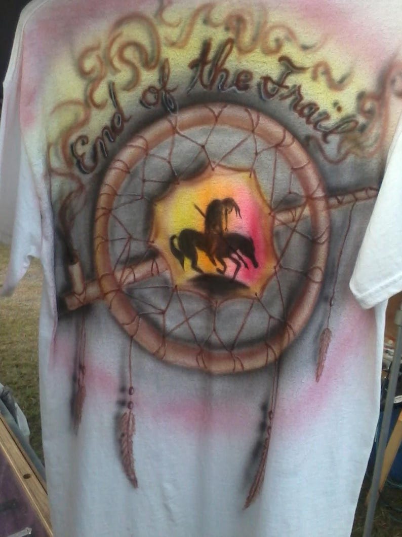 May include: White t-shirt with an airbrushed design featuring a dreamcatcher, a Native American figure on horseback, and the text "End of the Trail". The design incorporates shades of brown, pink, yellow, and black.