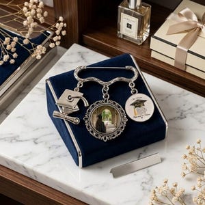 May include: A silver-toned graduation brooch with three charms: a mortarboard, a framed photo, and a circular charm with text. The brooch is displayed on a navy blue velvet box, with a marble surface in the background.