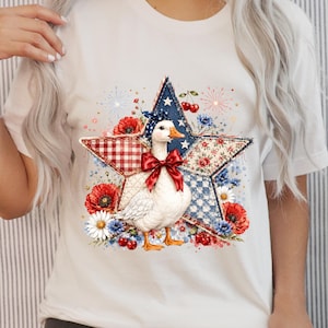 Può includere: T-shirt bianco con un design patriottico. Un'oca bianca con un papillon rosso è centrata su un design patchwork a forma di stella con motivi rossi, bianchi e blu e accenti floreali. Il design evoca il tema del 4 luglio.