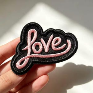 May include: An embroidered patch featuring the word love in light pink cursive script. The lettering is set against a black background with a thick, contoured black border that follows the shape of the text, creating a bold, high-contrast design.