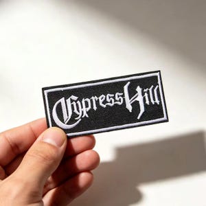 May include: A rectangular embroidered patch featuring a black background with a white border and the text Cypress Hill written in a stylised white gothic font. The patch has a textured fabric surface and is designed for application to clothing.