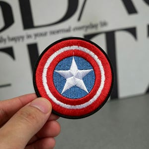 Op de afbeelding: Ronde geborduurde patch met een blauwe cirkel in het midden met een witte ster, omringd door rode en witte concentrische cirkels. De patch heeft een zwarte rand, waarschijnlijk een item met een superheldenthema.