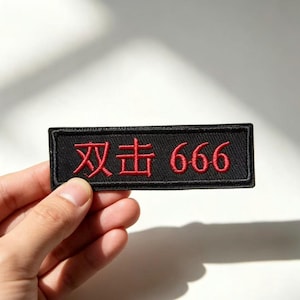Puede incluir: Un parche rectangular negro bordado con hilo rojo. El diseño incluye dos caracteres chinos seguidos del número 666. El parche tiene un borde cosido limpio y se presenta en una orientación horizontal con texto rojo claramente visible.