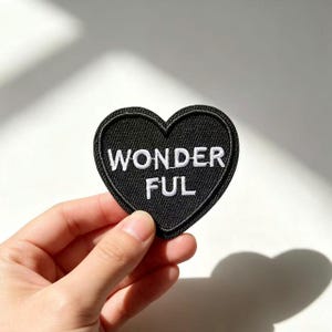 Pode incluir: Um emblema bordado em forma de coração com fundo preto e borda costurada em preto. A palavra WONDER está bordada em letras maiúsculas brancas na linha superior, e a palavra FUL está bordada em letras maiúsculas brancas abaixo.