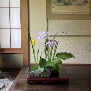 以下が含まれることがあります： 薄紫と白のアイリスの花2輪と小さな黄色のアイリスの花1輪、長い緑の葉と幅の広い葉を組み合わせたフラワーアレンジメント。質感のある長方形の灰色の陶器製容器に入れられ、木製の台座の上に置かれています。