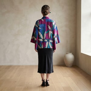 以下が含まれることがあります： クリーム、ブルー、マゼンタの色合いで大きな様式化された葉の鮮やかな模様が特徴の半袖ジャケット。シンプルな黒のミディ丈スカートの上に着用されています。ジャケットは構造的な襟と、まっすぐでゆったりとしたシルエットが特徴です。