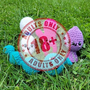 May include: A collection of handmade crochet toys in various colors, including white, pink, teal, and purple, resting on a bed of green grass. The image is overlaid with a red stamp that reads "ADULTS ONLY 18+".