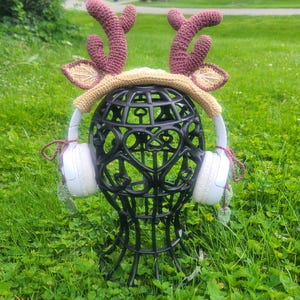 May include: A whimsical deer antler headband with crochet antlers in shades of burgundy and tan, paired with white headphones. The headband is displayed on a black decorative head form, set against a backdrop of green grass.