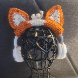 May include: A handmade orange and white crochet fox ear headband. The headband is placed on a black decorative head-shaped stand, with white headphones on either side. The ears are detailed with white inner ear accents.