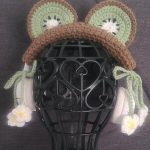May include: A whimsical, handmade crochet hat resembling a kiwi fruit. The hat features green and brown yarn, with white and yellow flower accents. The hat is displayed on a black wire form, showcasing its unique design.