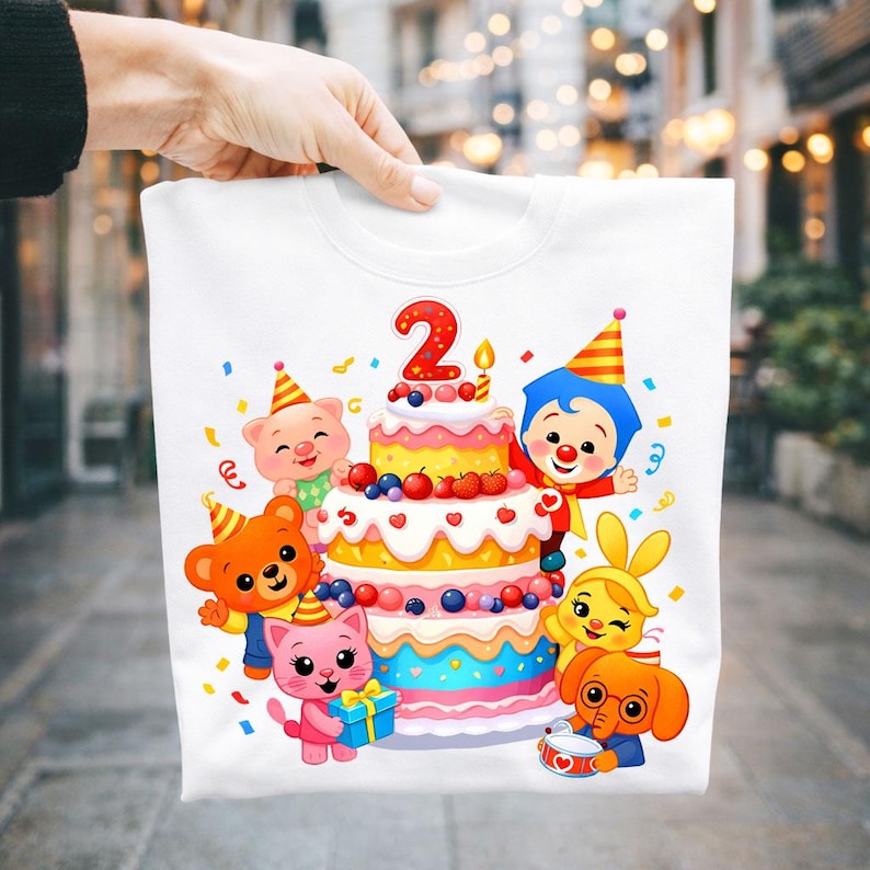 May include: A white t-shirt featuring a colorful graphic of a three-tiered birthday cake topped with a red number two candle. Surrounding the cake are cartoon characters including a pig, bear, cat, clown, rabbit, and elephant with confetti.