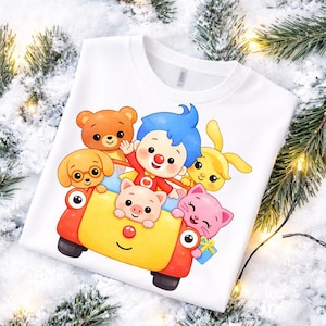 May include: A white t-shirt featuring a colorful graphic of a cartoon clown with blue hair and a group of animal friends, including a brown bear, a yellow dog, a pink pig, and a pink cat, all riding in a bright red and yellow toy car.