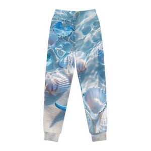 May include: Jogger pants with a beach-themed design. The pants feature a print of seashells, starfish, and sand under clear water. The color palette includes shades of blue, white, and beige. The cuffs and waistband are gray.