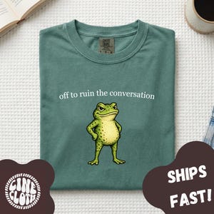 Könnte beinhalten: Ein salbeigrünes T-Shirt mit einer Grafik eines stehenden Frosches mit strengem Gesichtsausdruck und dem weißen Text "off to ruin the conversation" über dem Frosch. Das T-Shirt hat einen Rundhalsausschnitt und ein Comfort Colors Markenetikett.