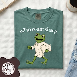 Peut inclure: Un t-shirt vert sauge avec une illustration d'une grenouille verte portant un pyjama blanc à motifs de lunes et d'étoiles, tenant un oreiller blanc. Le texte "off to count sheep" est imprimé en blanc au-dessus. Étiquette Comfort Colors visible.