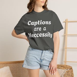 May include: A dark gray t-shirt with the text "Captions are a Necessity" in white. The model is wearing denim shorts. The shirt is a crew neck and has short sleeves.