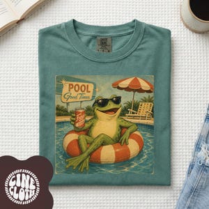 May include: A teal t-shirt featuring a graphic of a green frog wearing black sunglasses, relaxing in a red and white striped pool float while holding a can of soda. The background shows a swimming pool, a lounge chair, and a red and white umbrella.