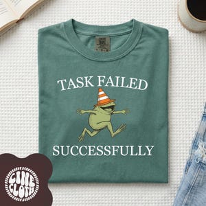 Puede incluir: Camiseta verde con la frase "TASK FAILED SUCCESSFULLY" y una rana de dibujos animados con un sombrero de cono de tráfico. La camiseta es de cuello redondo.