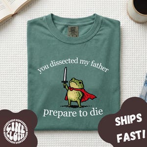 Può includere: Una t-shirt verde salvia con la grafica di una rana verde che indossa un mantello rosso e impugna una spada. Il testo sopra la rana recita "you dissected my father" e il testo sottostante "prepare to die". La maglia ha un girocollo.
