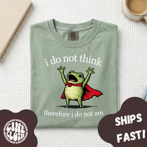 May include: A sage green t-shirt featuring a graphic of a green frog wearing a red cape with its arms raised. The text above the frog reads "i do not think" and the text below reads "therefore i do not am" in white, lowercase lettering.