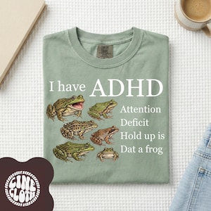 May include: Sage green t-shirt with the text "I have ADHD" and a graphic of six different frog illustrations. Additional text reads "Attention Deficit Hold up is Dat a frog".