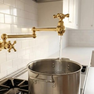 Op de afbeelding: Een goudkleurige, aan de muur gemonteerde potvuller met een kruisvormige handgreep is uitgeschoven boven een roestvrijstalen pot op een gasfornuis. Water stroomt uit de kraan in de pot. De keuken heeft witte tegels en kasten.
