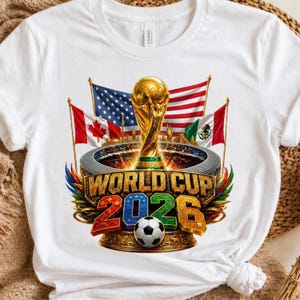 K&ouml;nnte beinhalten: Wei&szlig;es T-Shirt mit einem farbenfrohen Grafikdesign f&uuml;r die Weltmeisterschaft 2026. Das Design zeigt die Weltpokal-Troph&auml;e, die Flaggen der USA, Kanadas und Mexikos sowie den Text "WORLD CUP 2026" mit einem Fu&szlig;ball.