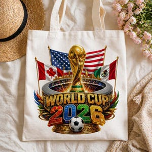 K&ouml;nnte beinhalten: Eine wei&szlig;e Tragetasche mit einem goldenen World Cup-Pokal und dem Text "WORLD CUP 2026". Das Design umfasst die Flaggen der Vereinigten Staaten, Kanadas und Mexikos. Ein Fu&szlig;ball ist ebenfalls abgebildet.
