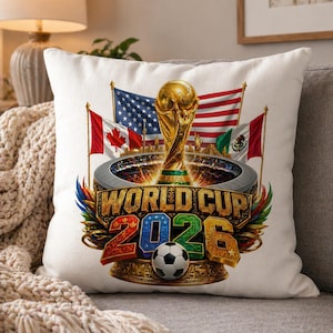 K&ouml;nnte beinhalten: Quadratisches wei&szlig;es Kissen mit einem farbenfrohen Grafikdesign f&uuml;r die Weltmeisterschaft 2026. Das Design zeigt die FIFA-Weltmeisterschaftstroph&auml;e, Flaggen der USA, Kanadas und Mexikos sowie den Text "WORLD CUP 2026" mit einem Fu&szlig;ball.
