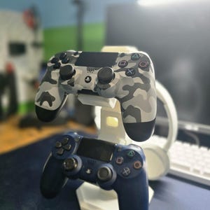 May include: A white stand holding two game controllers. The top controller has a gray and white camouflage pattern. The bottom controller is solid navy blue. Both controllers have black buttons and directional pads.