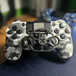 May include: A camouflage-patterned video game controller with a black accessory attached. The controller has a black touchpad, buttons with colored symbols, and a black accessory with a circular design. A blue controller is visible in the background.