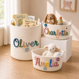 May include: Three off-white woven rope storage baskets featuring colorful embroidered names and small flower accents. The largest basket displays the name Oliver, the medium basket says Charlotte, and the smallest basket features the name Amelia.