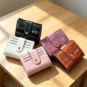 May include: Five small bi-fold wallets with a crocodile-embossed texture, each featuring a snap-button closure and gold-tone hardware. The wallets are available in black, white, pink, mauve, and brown, with gold-lettered monograms WT, QR, and VH.